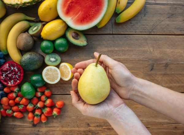 Hands holding a pear surrounded by a colorful variety of fruits on a wooden surface.