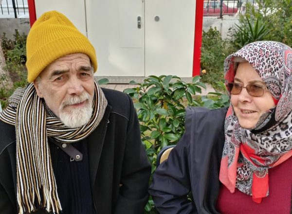 Elderly man and woman sitting outdoors, wearing winter attire with scarves. Bright day scene.